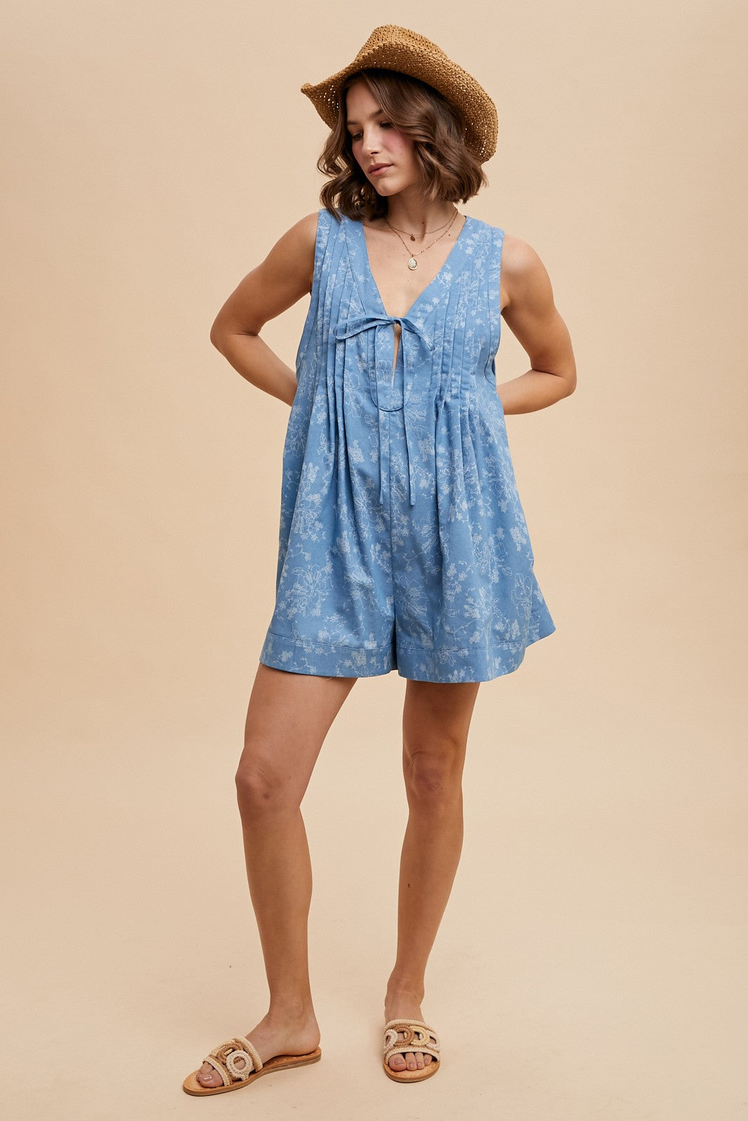 woman wearing a light blue floral romper with a v-neckline and relaxed fit, accessorized with a straw hat