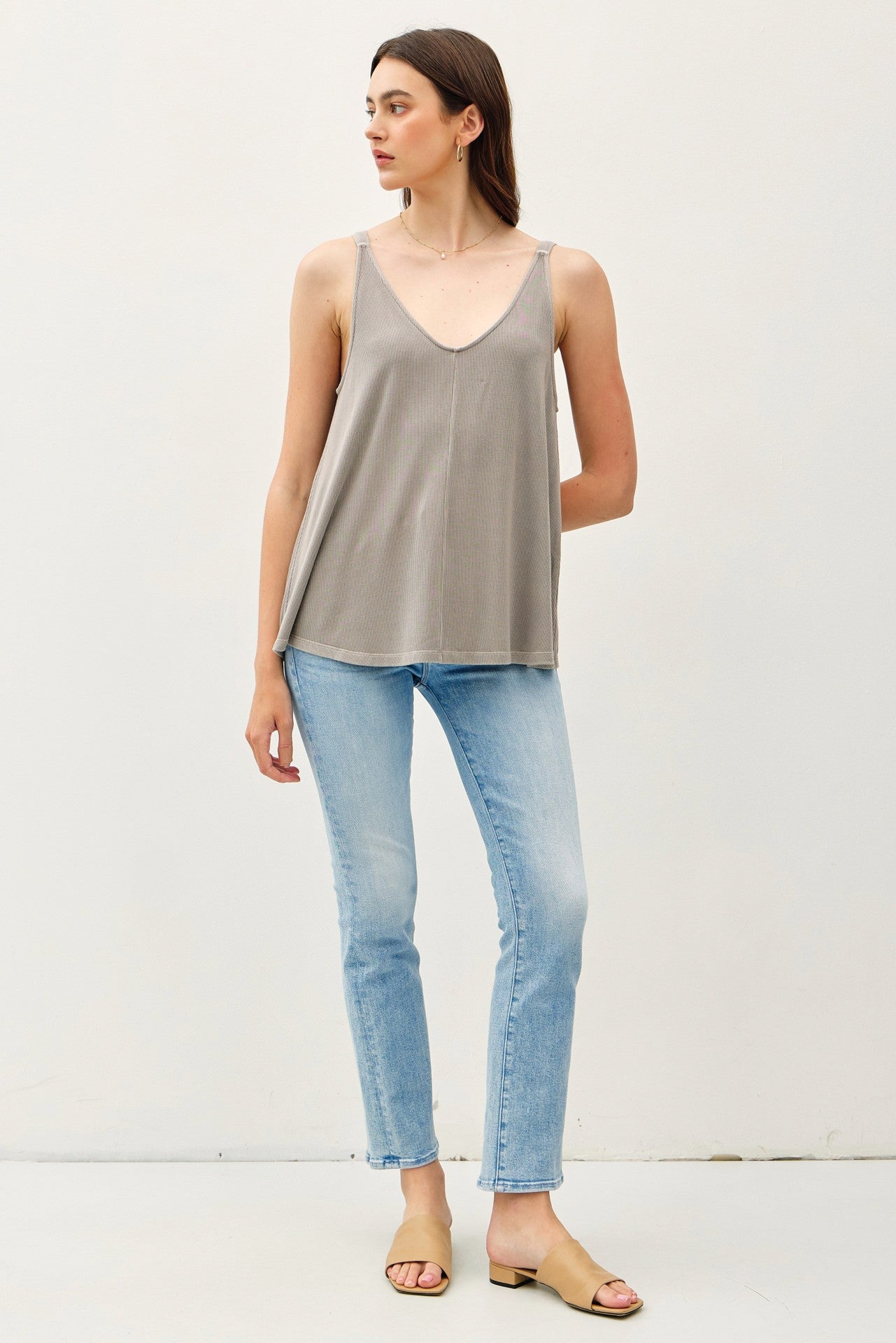 woman wearing a light gray spaghetti strap tank top with a v-neck and blue skinny jeans, standing against a plain background