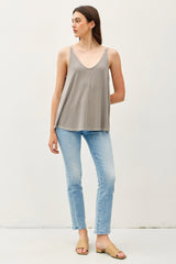 woman wearing a light gray spaghetti strap tank top with a v-neck and blue skinny jeans, standing against a plain background
