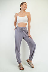 woman wearing a white cropped tank top and light gray jogger pants, standing with hands in pockets and smiling