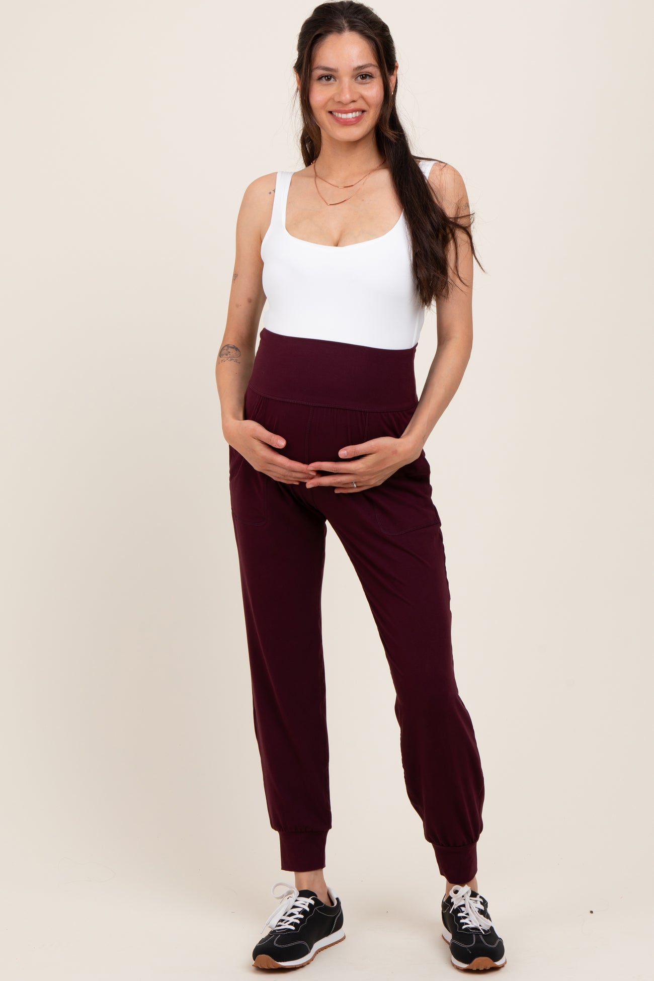 woman wearing a white maternity bodysuit and maroon maternity joggers, gently holding her baby bump