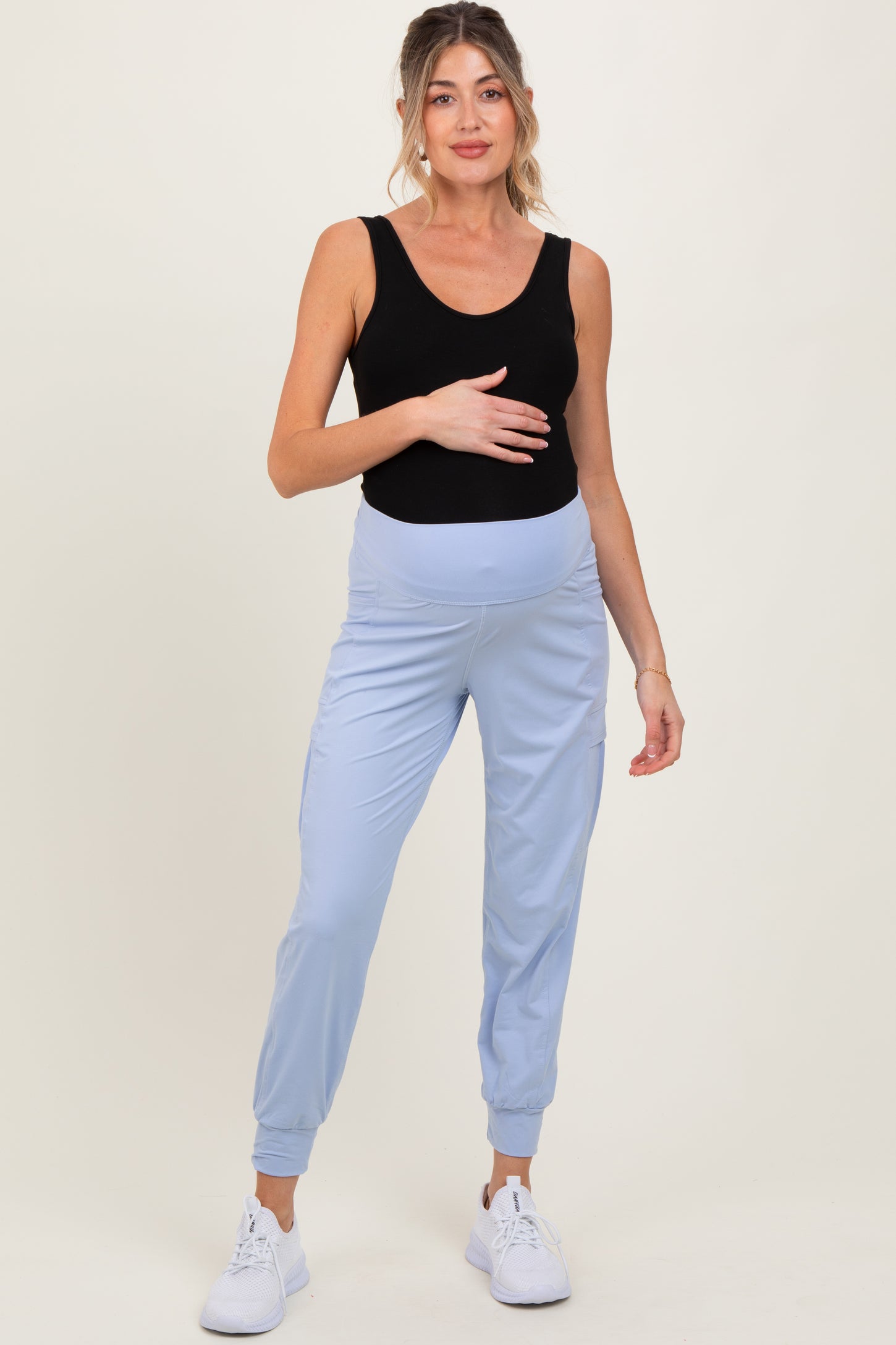 woman wearing a black tank top and light blue maternity joggers, showing her baby bump while posing