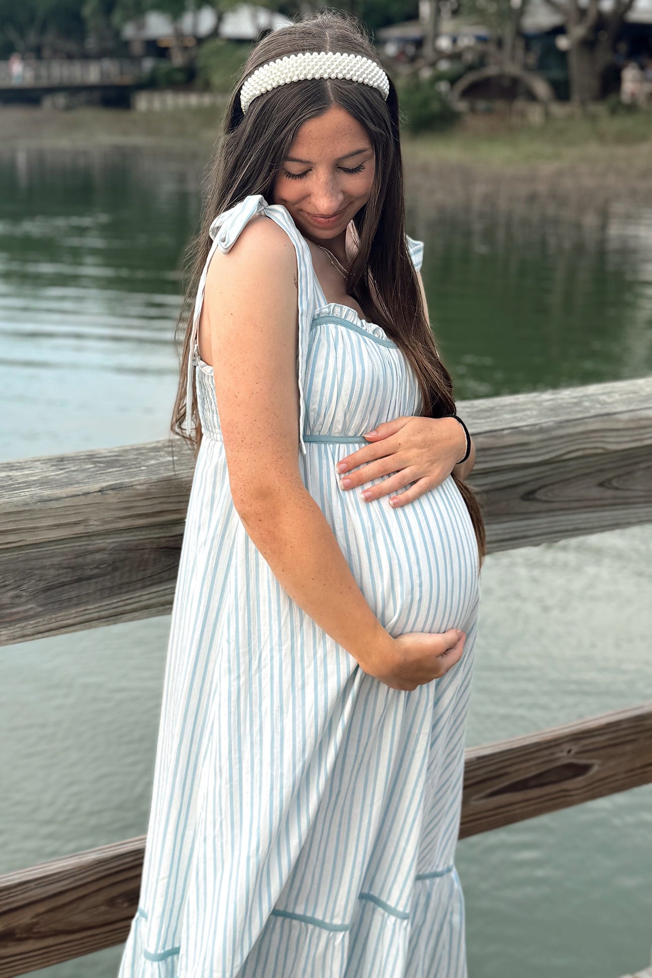 woman wearing a striped maternity dress with tie straps and cradling her baby bump by the water