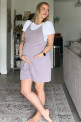 woman wearing a lavender ribbed maternity overall dress and a white t-shirt, holding her baby bump in a cozy home setting