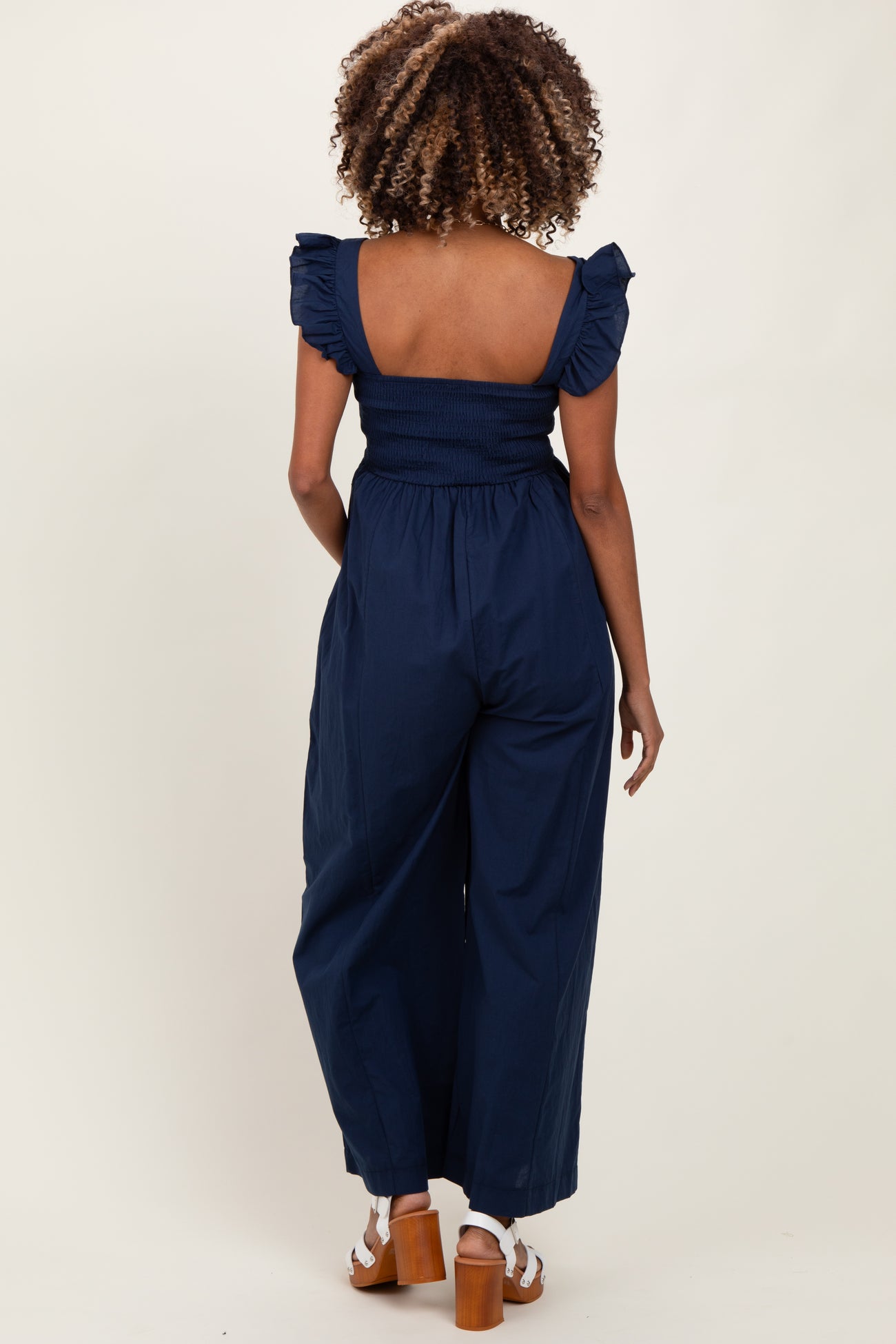 woman wearing a navy blue maternity jumpsuit with smocked bodice and ruffle sleeves, seen from the back