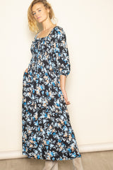 woman wearing a black floral maxi dress with three-quarter sleeves and gathered waist, posing with one hand in pocket