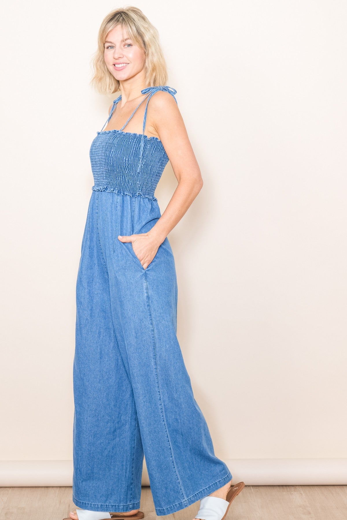 woman wearing a blue denim smocked wide-leg jumpsuit with adjustable tie straps and side pockets