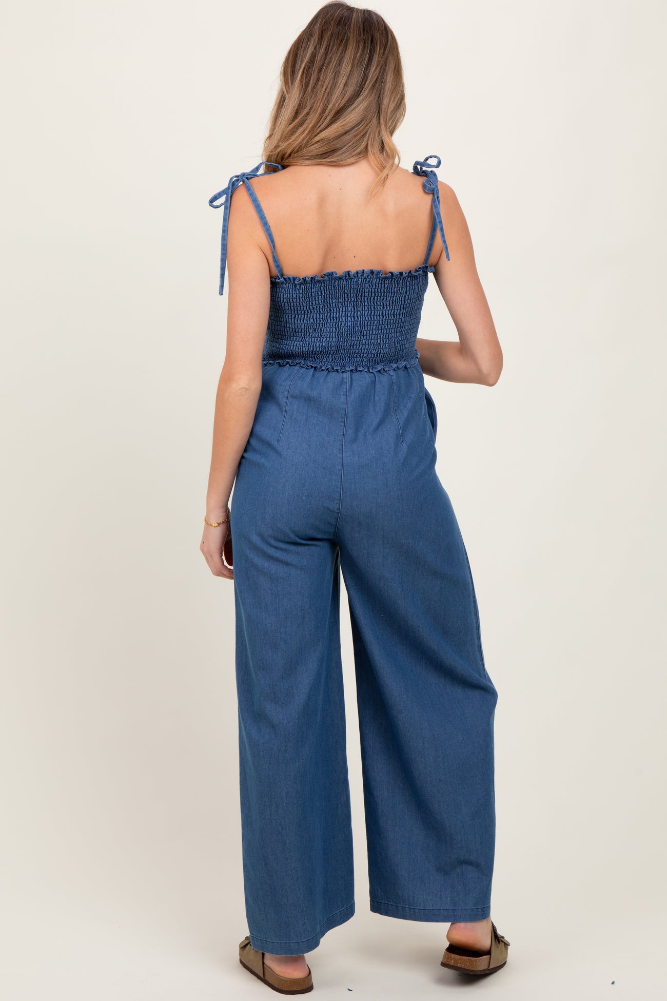 woman wearing a blue denim maternity jumpsuit with smocked bodice and wide leg pants, viewed from the back