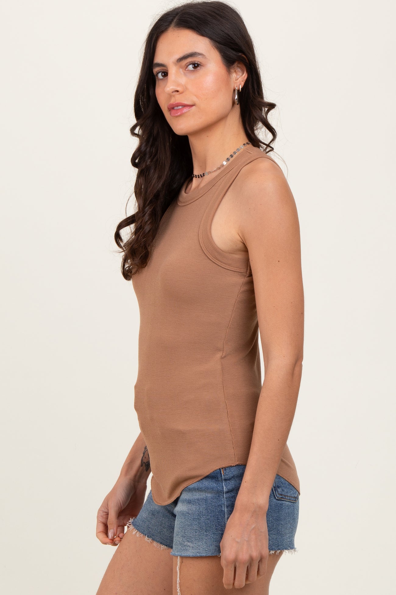 woman wearing a brown ribbed tank top and denim shorts, standing sideways with a relaxed expression
