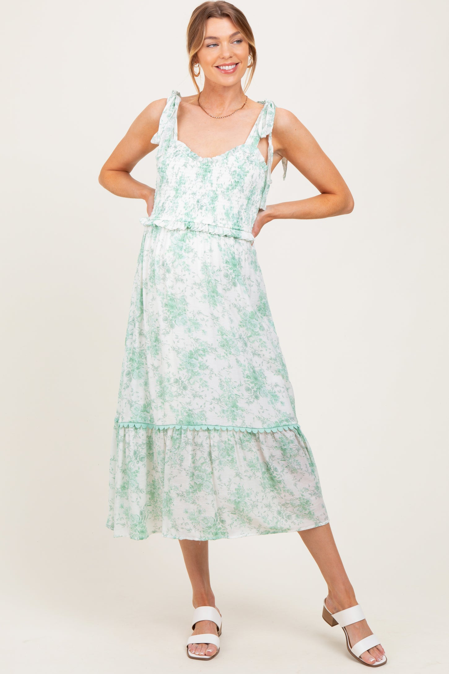 woman wearing a green floral print maternity dress with ruffled straps and a tiered skirt, smiling against a neutral background