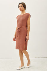 woman wearing a rust colored knit dress with a belted waist and side slits, styled with flat white shoes