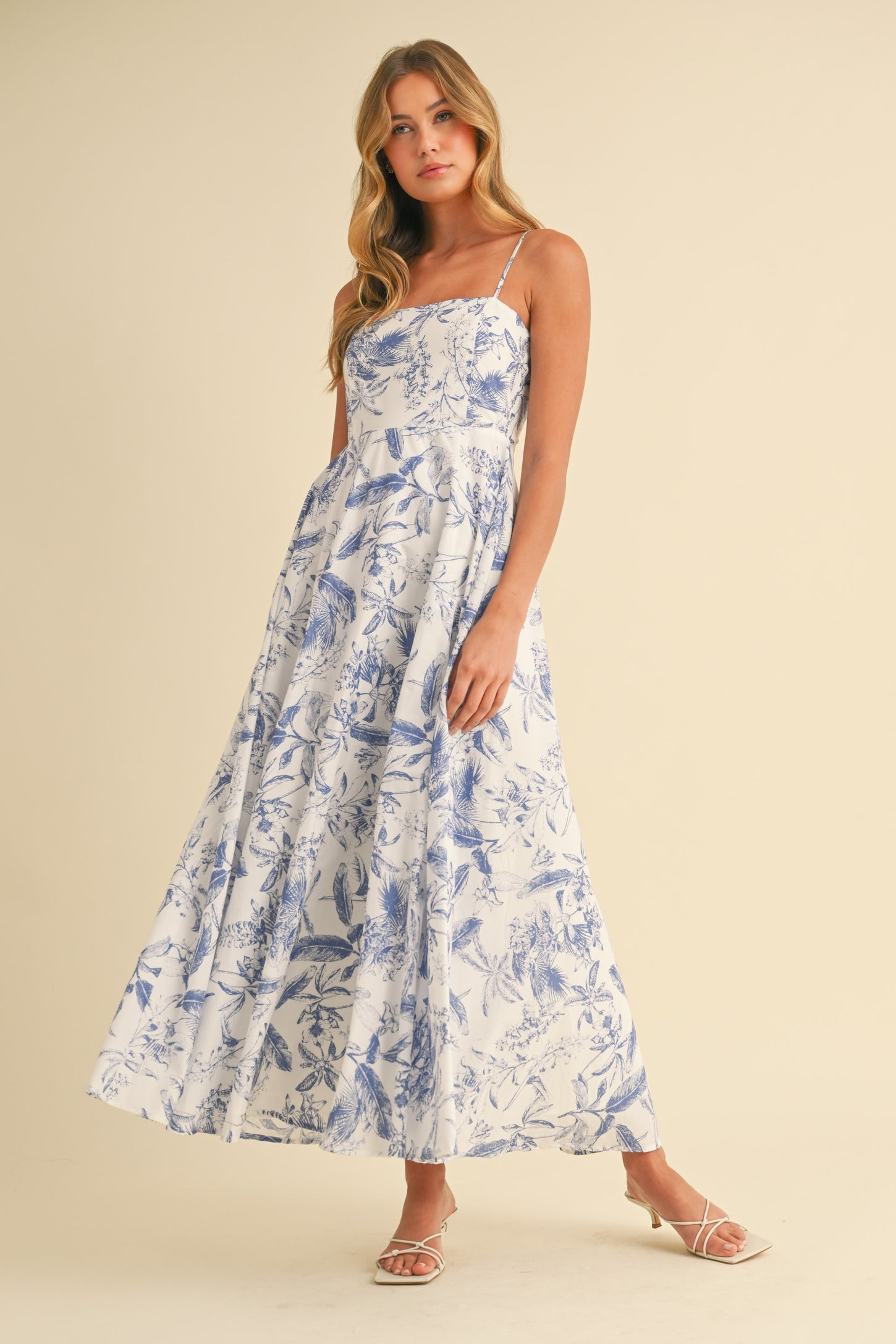 woman wearing a white floral maxi dress with blue patterns and thin straps, standing against a light background