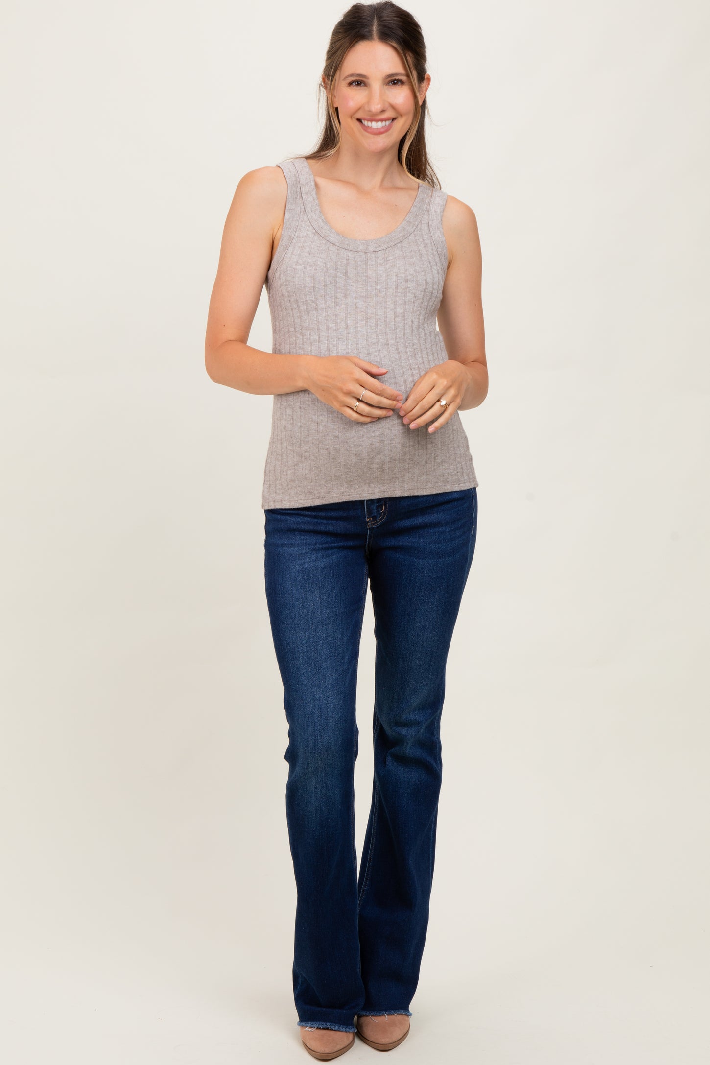 woman wearing a light gray ribbed maternity tank top and blue flared maternity jeans, smiling and showing her baby bump