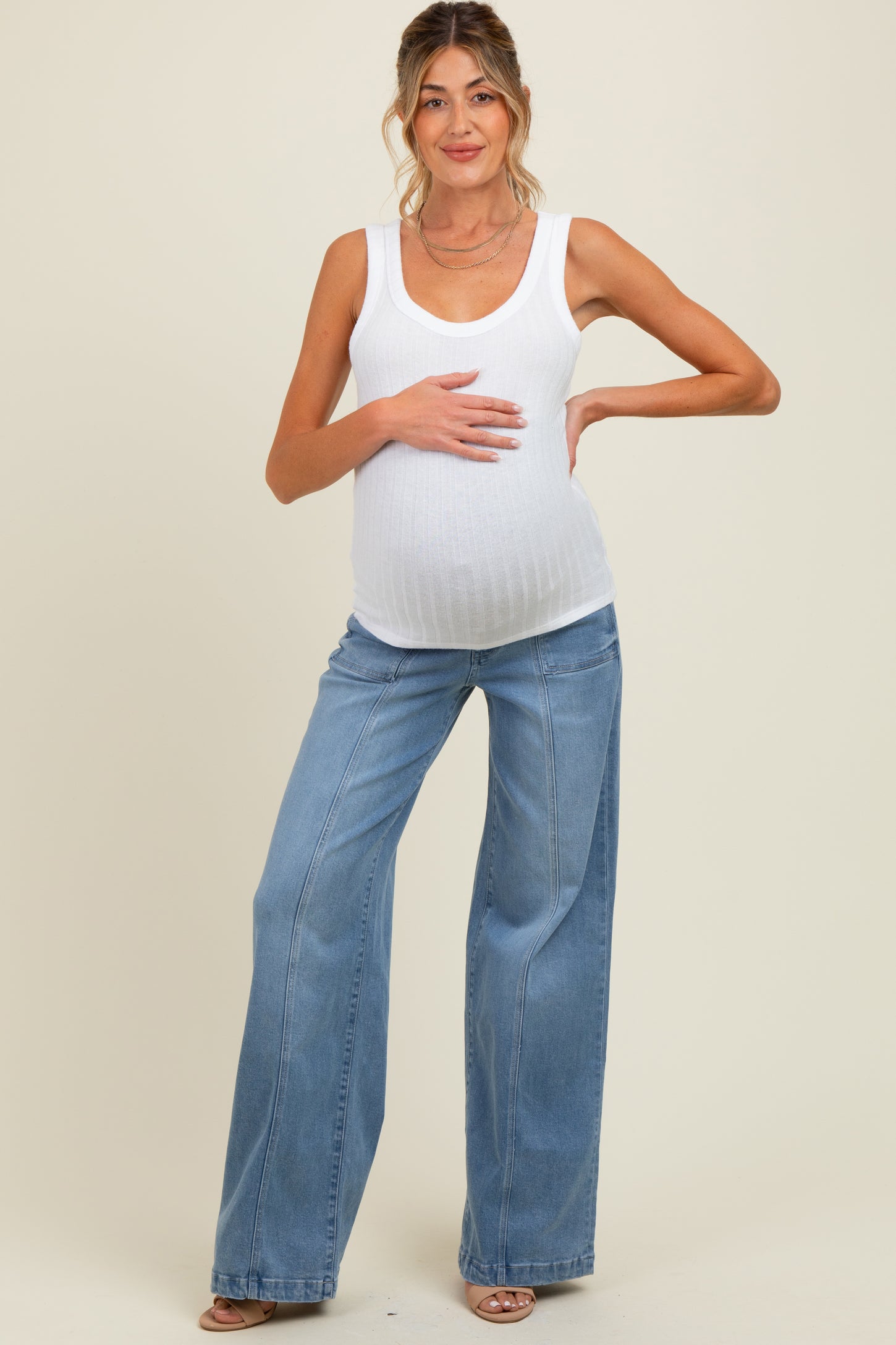 woman wearing a white ribbed maternity tank top and blue wide-leg maternity jeans, posing with her baby bump visible