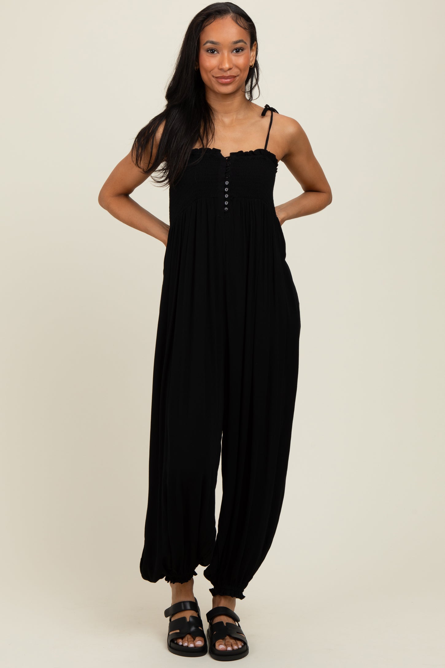 woman wearing a black smocked jumpsuit with adjustable straps and button detail, standing confidently with hands on hips