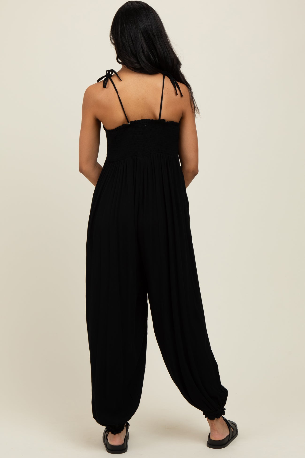 woman wearing a black sleeveless jumpsuit with smocked bodice and adjustable tie straps, viewed from the back