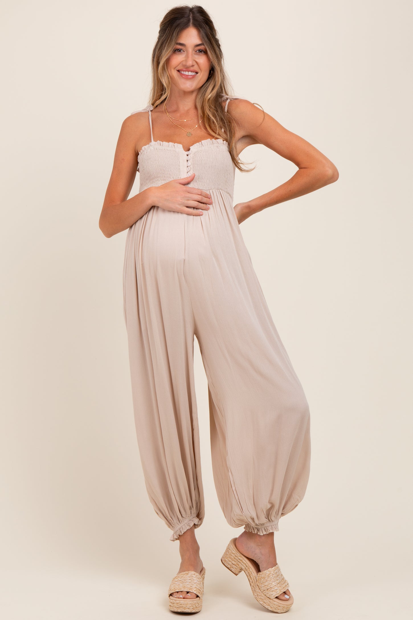 woman wearing a beige maternity jumpsuit with a smocked bodice and balloon legs, showing her baby bump
