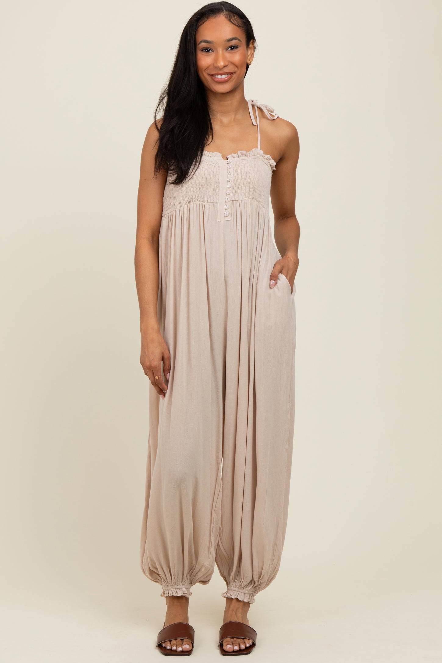 woman wearing a beige sleeveless jumpsuit with a smocked bodice and balloon legs, standing with hands in pockets