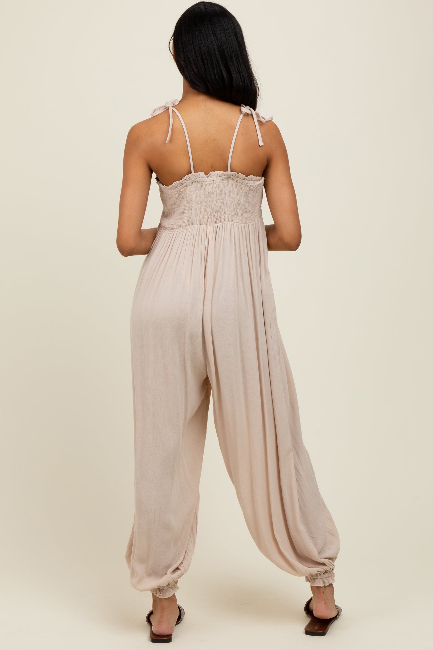 woman wearing a beige sleeveless jumpsuit with adjustable shoulder straps and a smocked bodice, viewed from the back