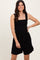 woman wearing a black ribbed mini dress with a square neckline and smocked bodice, smiling and posing with one hand on her hip