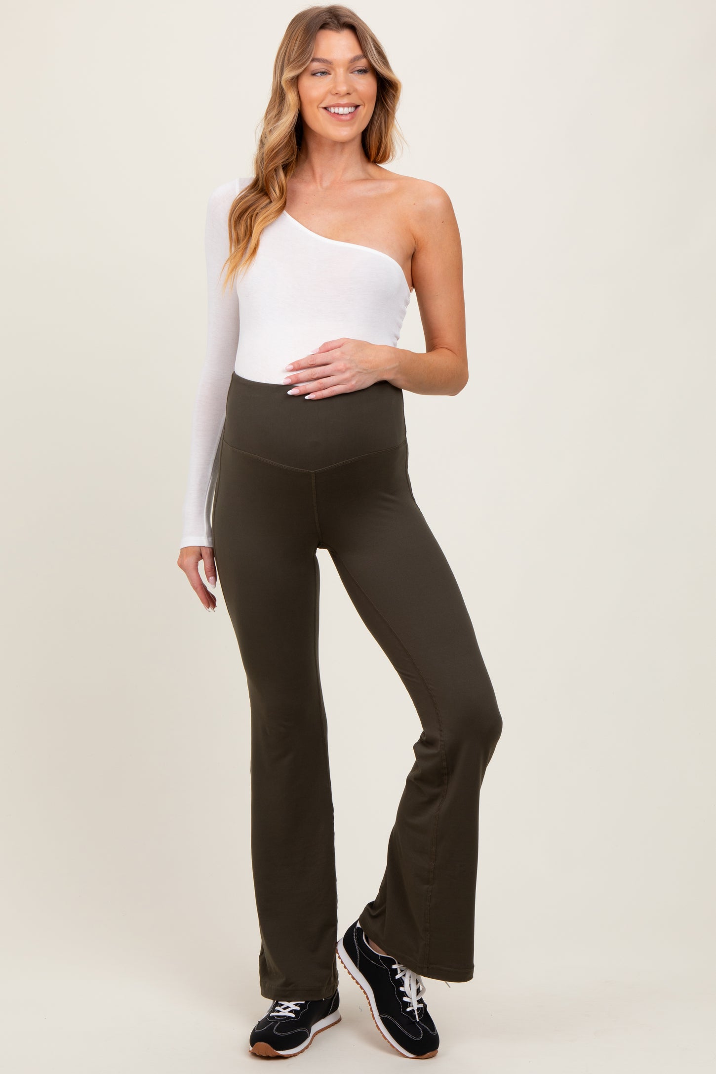 woman wearing a white one-shoulder top and olive green maternity flared leggings, smiling, with a baby bump visible
