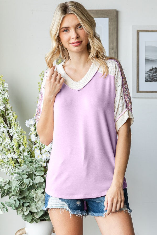 woman wearing a lilac v-neck top with embellished short sleeves and denim shorts, standing in a bright room with plants