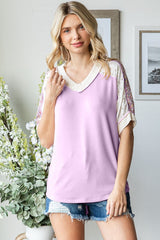 woman wearing a lilac v-neck top with embellished short sleeves and denim shorts, standing in a bright room with plants
