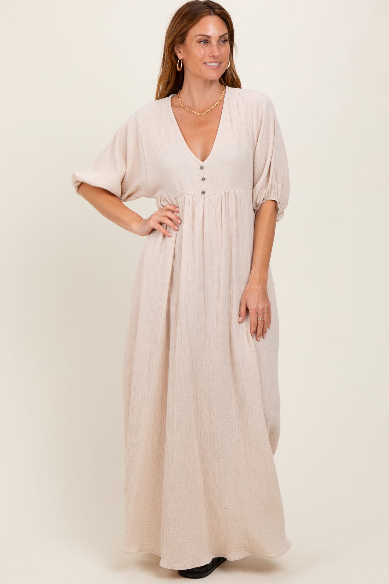woman wearing a light beige maxi dress with a deep v-neck and button details, standing with hands on hips