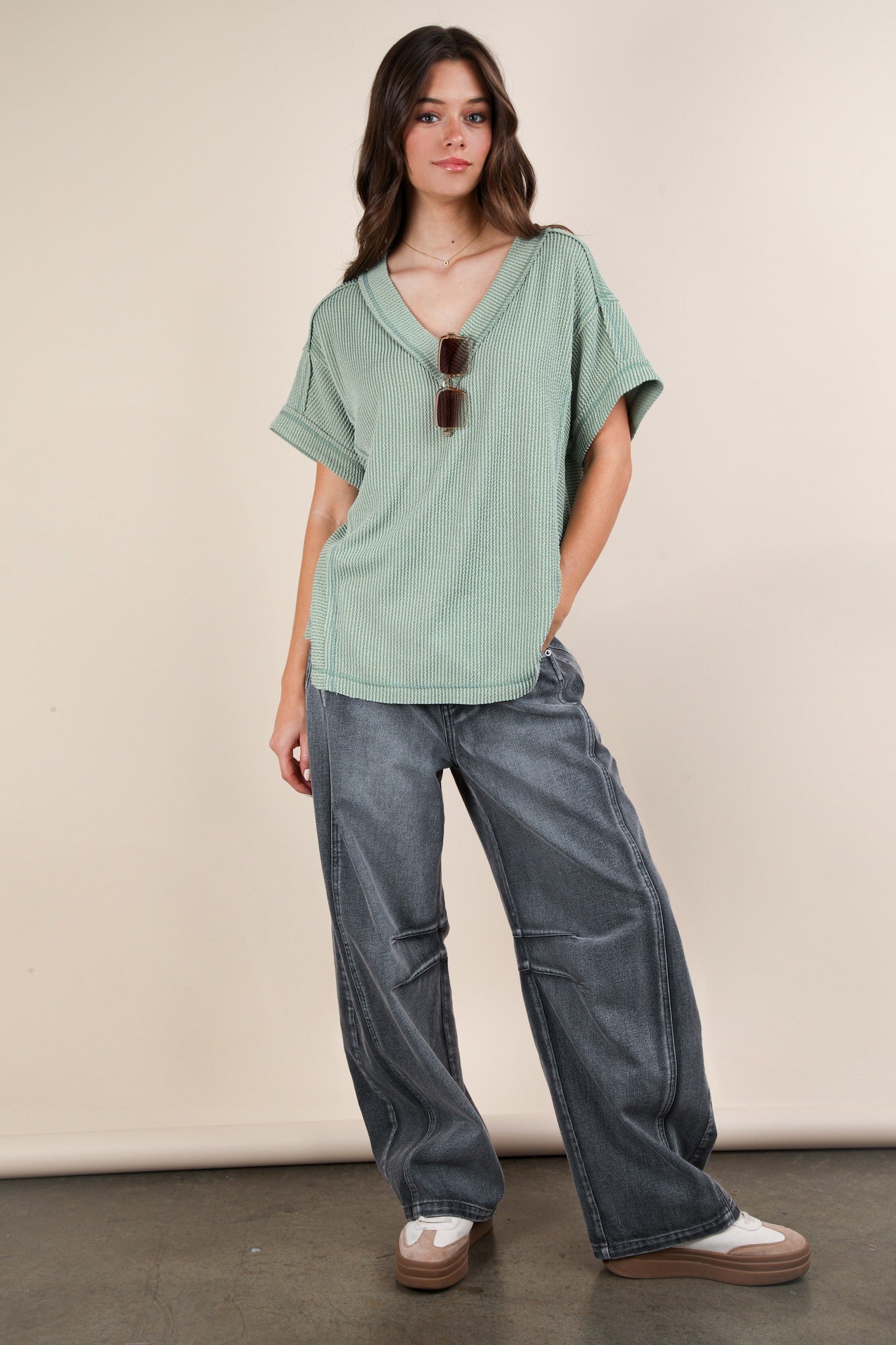 woman wearing a green striped oversized v-neck top and gray wide-leg jeans with sunglasses on her shirt