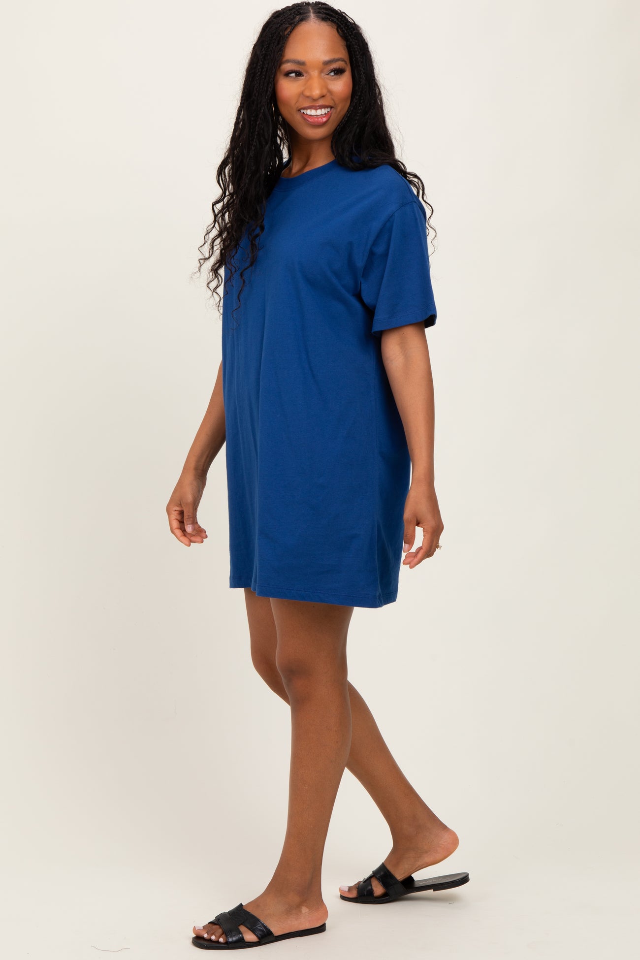woman wearing a short-sleeve blue t-shirt dress and black slide sandals, smiling while walking
