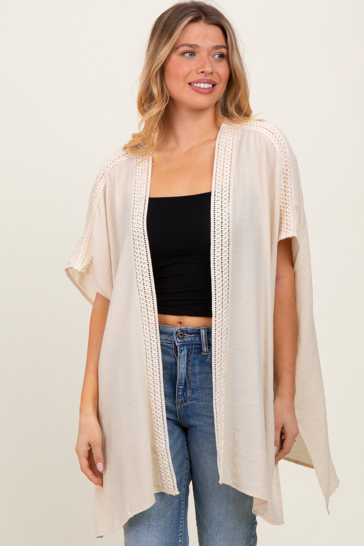 woman wearing a beige open-front kimono cardigan with lace details over a black crop top and blue jeans