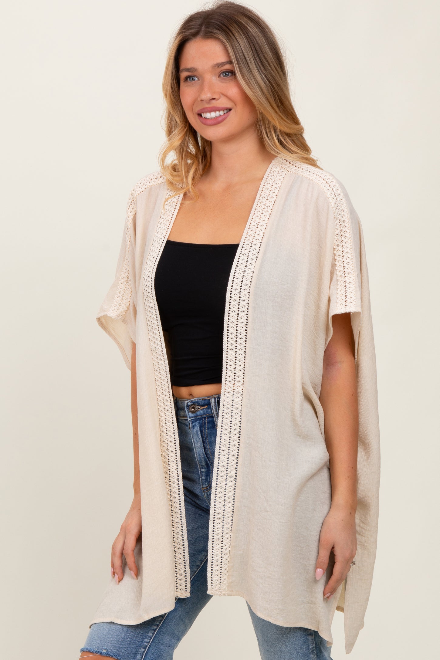 woman wearing a beige kimono style cardigan with crochet details over a black crop top and blue jeans