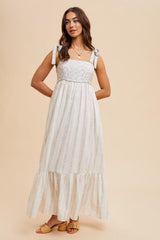 woman wearing a cream floral maxi dress with adjustable tie straps and a tiered skirt, paired with tan sandals