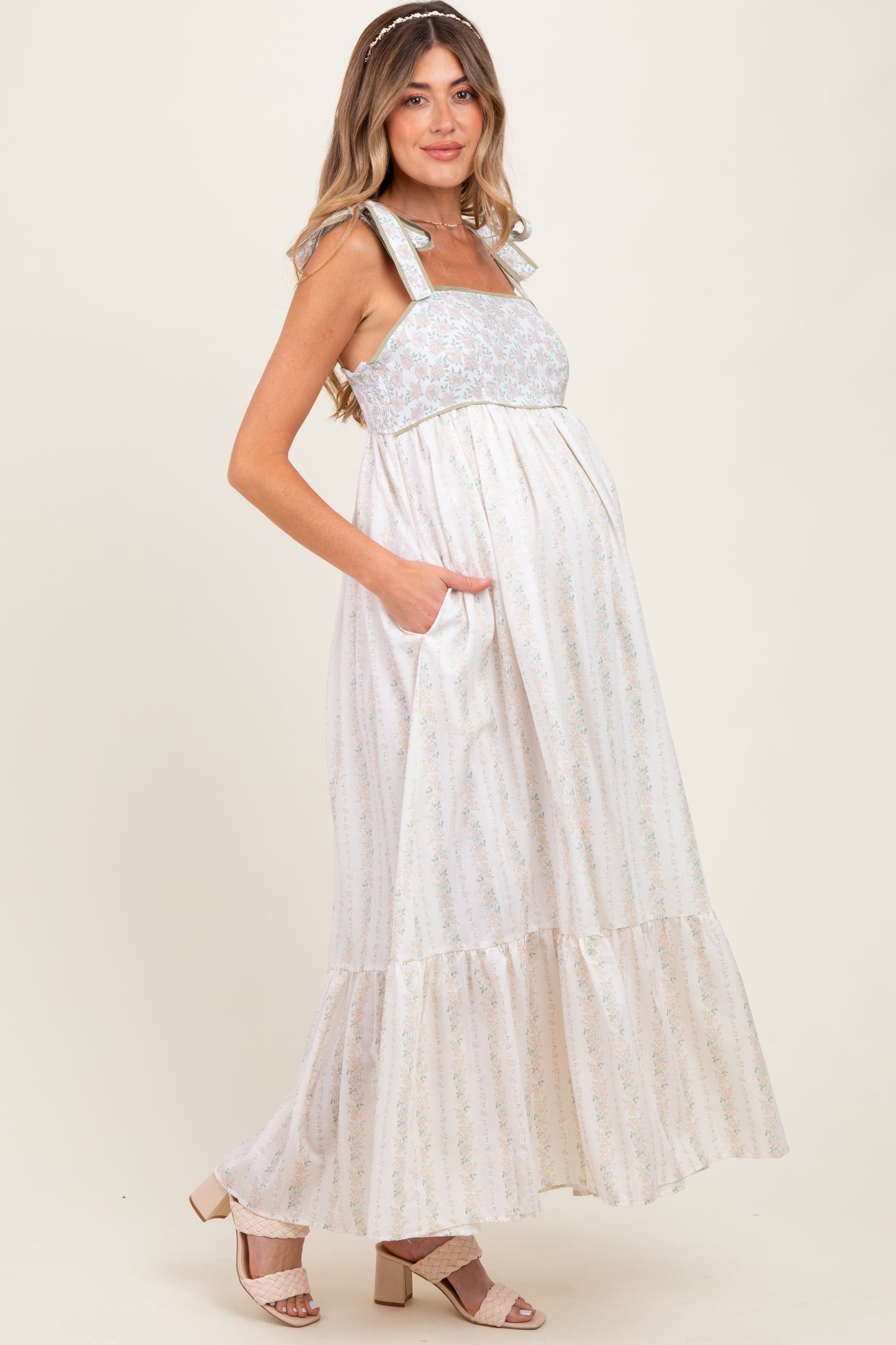 woman wearing a cream floral print maternity maxi dress with pockets and a ruffled hem, showcasing her baby bump