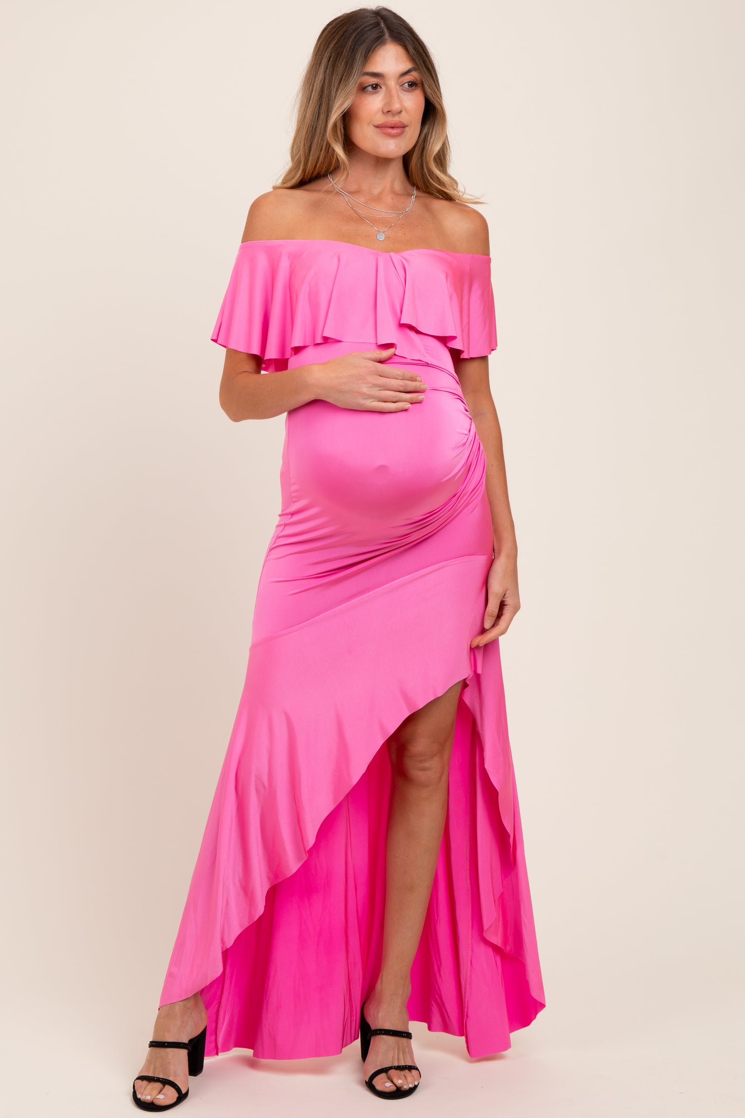woman wearing a hot pink off-the-shoulder maternity dress with a ruffled top and high-low hem, holding her baby bump