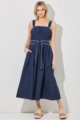 woman wearing a navy blue maxi dress with a drawstring waist and oversized straps, standing with pockets and open-toe sandals