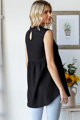 woman wearing a black sleeveless top with a keyhole back and peplum style, standing in a bright room with plants