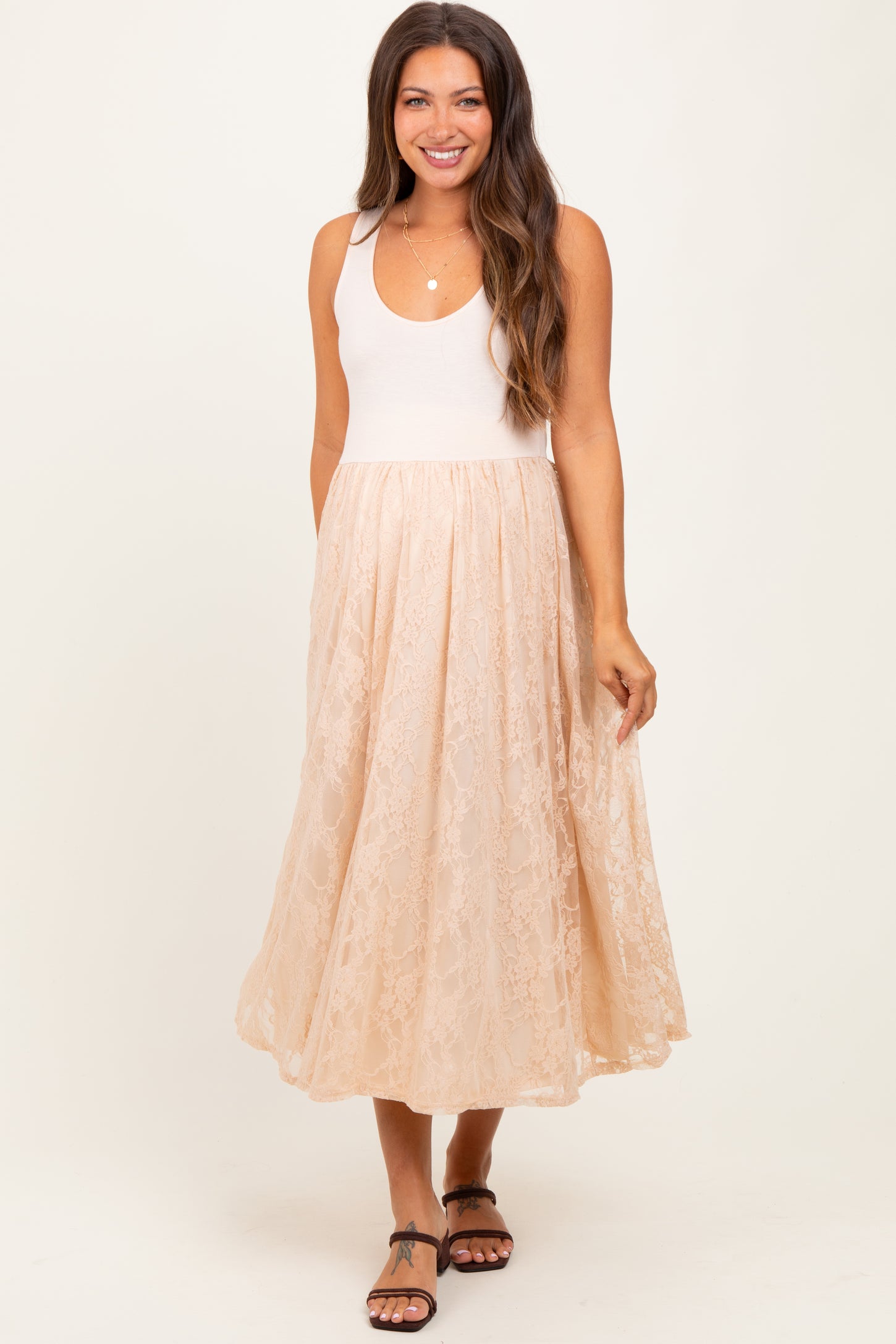 woman wearing a light pink maternity tank top with a flowy beige lace skirt and smiling at the camera