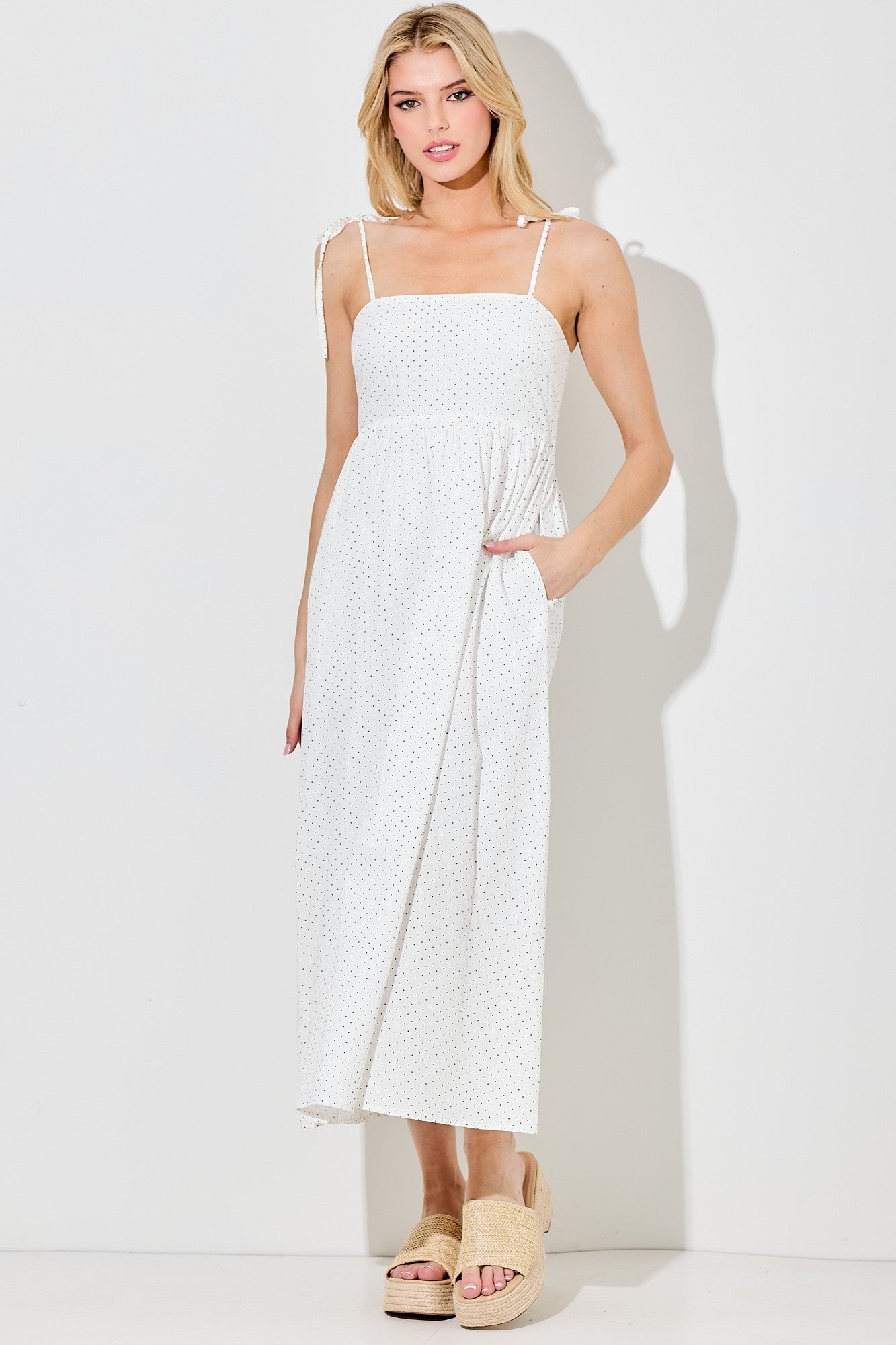woman wearing a white polka dot maxi dress with adjustable straps and pockets, styled with beige slide sandals