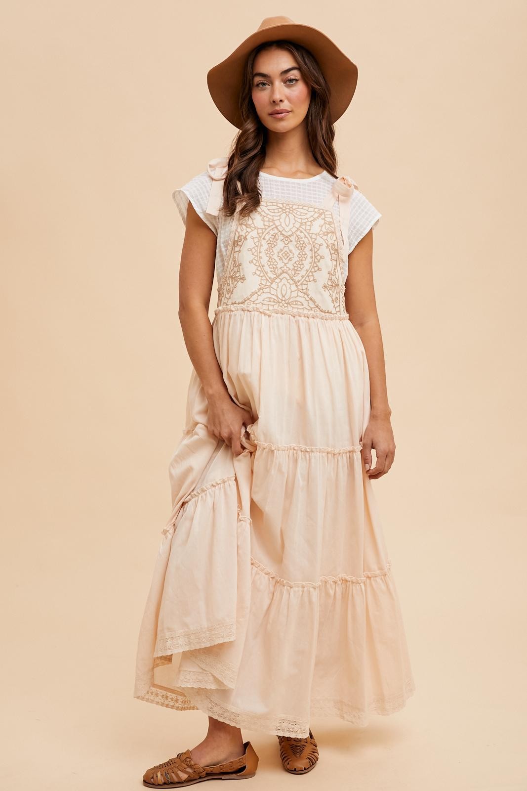 woman wearing a light peach maxi dress with crochet details and a white top, holding the dress and wearing a wide-brimmed hat