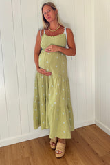 woman wearing a green maternity dress with tiered ruffles and white embroidery, holding her baby bump