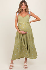 woman wearing a light green maternity dress with floral embroidery and ruffled tiers, holding her baby bump