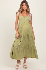woman wearing a light green maternity dress with tiered layers and blue floral embroidery, featuring shoulder ties and a baby bump