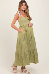 woman wearing a green maternity maxi dress with a smocked bodice, floral embroidery, and side pockets, holding her baby bump