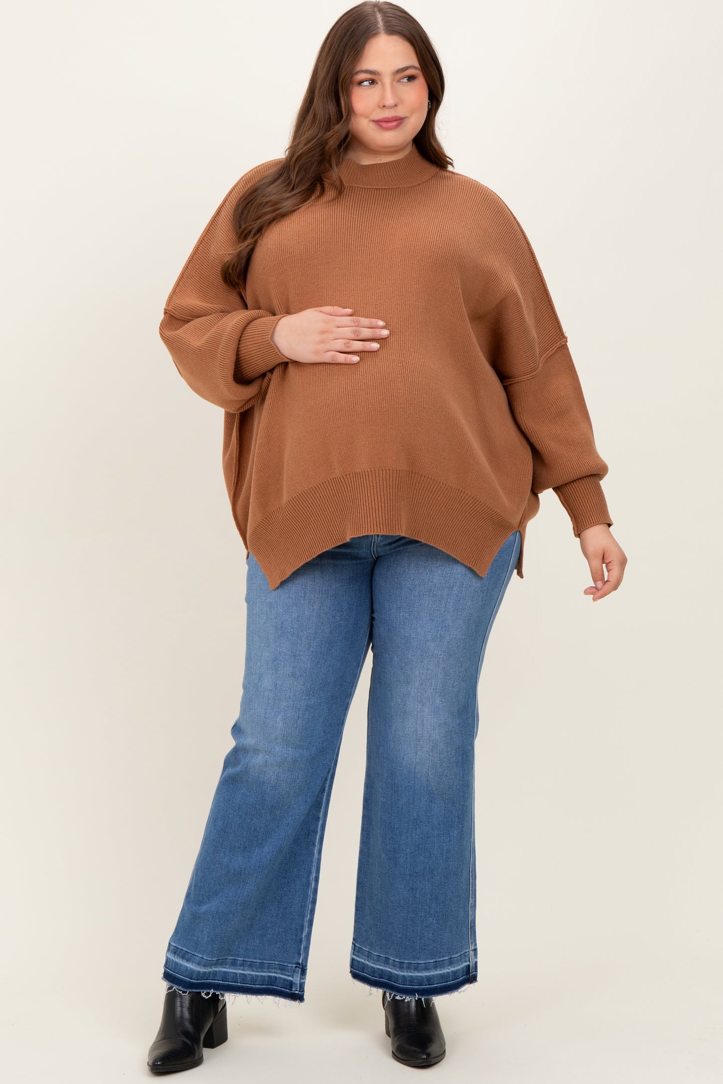woman wearing a brown oversized maternity sweater and blue flared maternity jeans, holding her baby bump