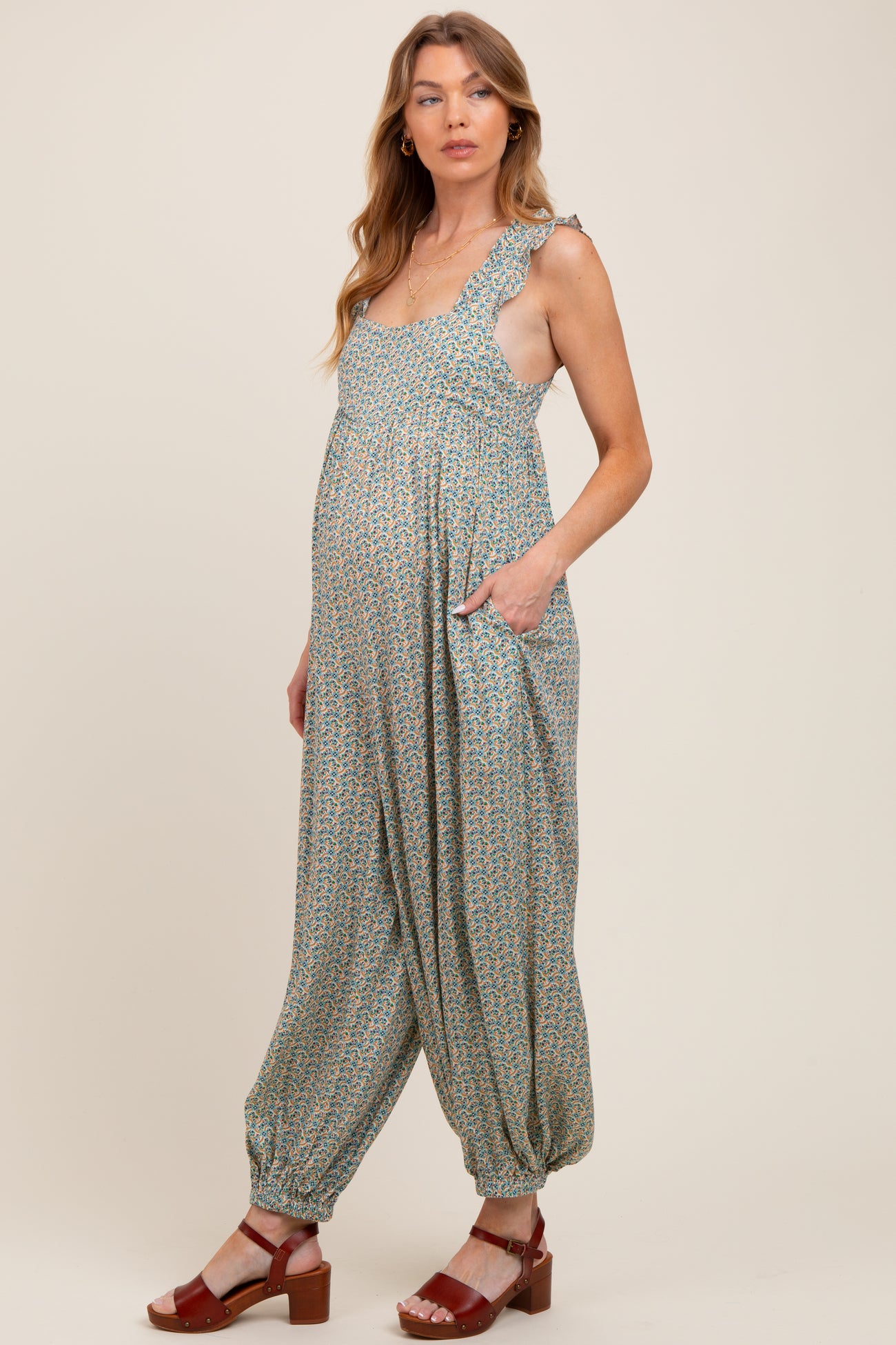 woman wearing a green floral maternity jumpsuit with ruffled straps and pockets, showing her baby bump