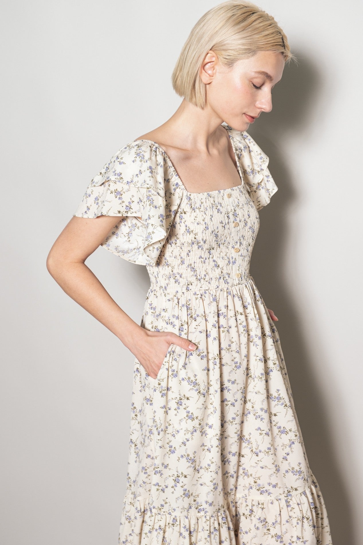 woman wearing a cream floral print maxi dress with smocked bodice and flutter sleeves, featuring side pockets