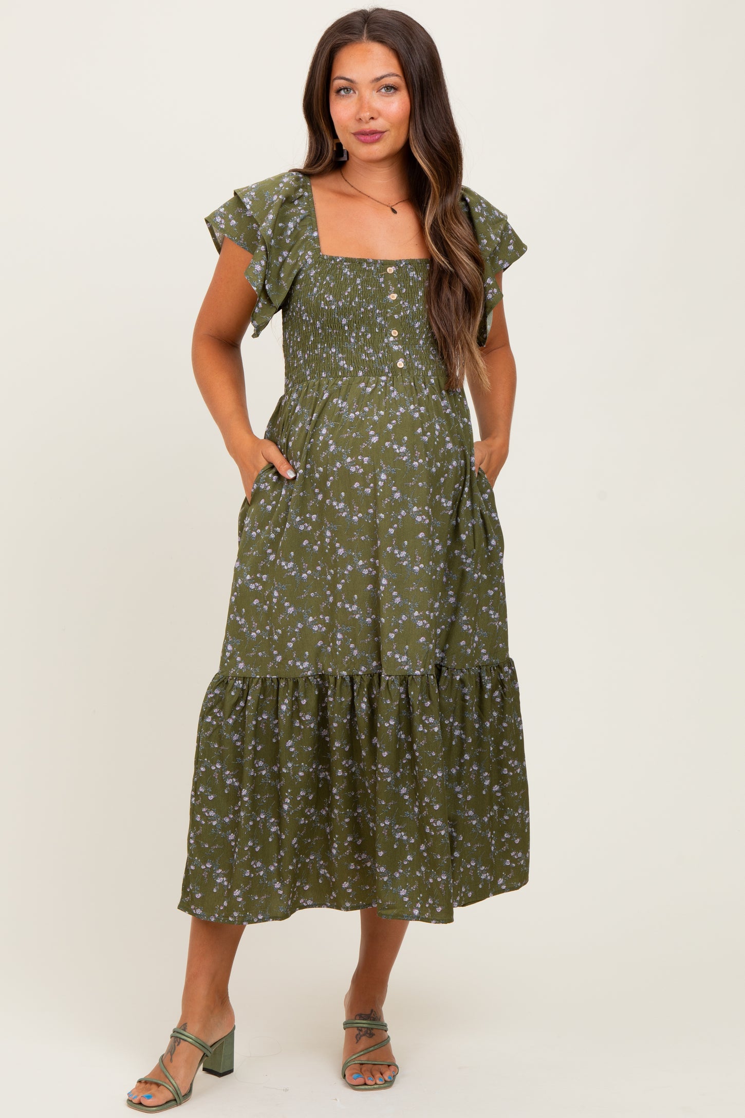 woman wearing a green floral print maternity dress with flutter sleeves and pockets, styled with sandals