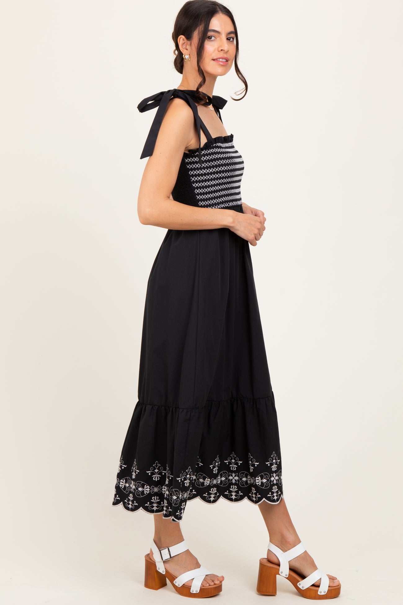 woman wearing a black maxi dress with white embroidery and tie straps, styled with white block-heeled sandals