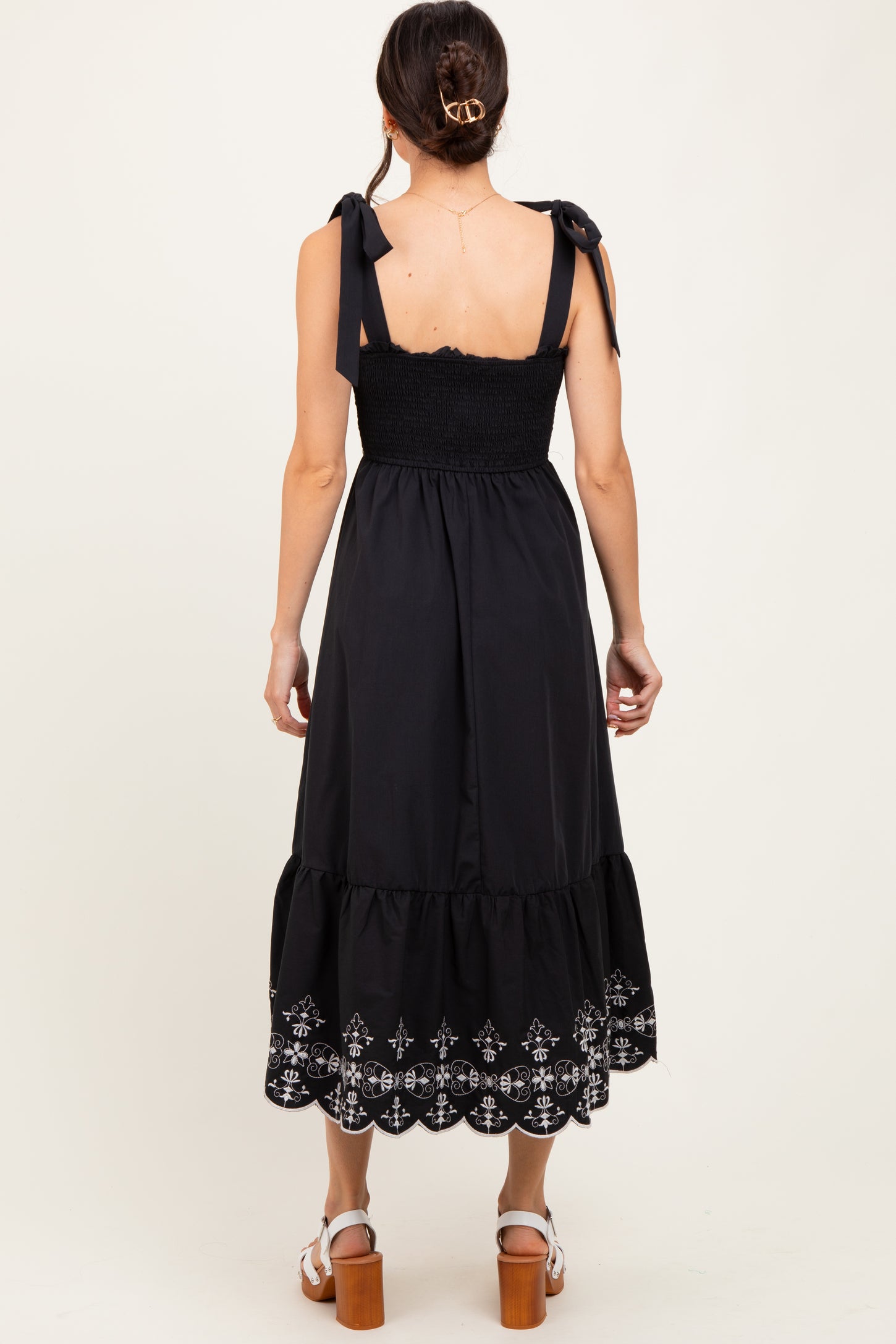 woman wearing a black maxi dress with smocked bodice, adjustable tie straps, and embroidered hem detail